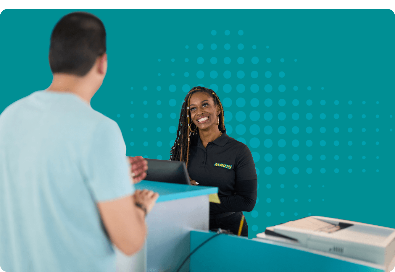 Smiling employee in black Mavis shirt assists customer at service desk with teal background and dotted pattern.